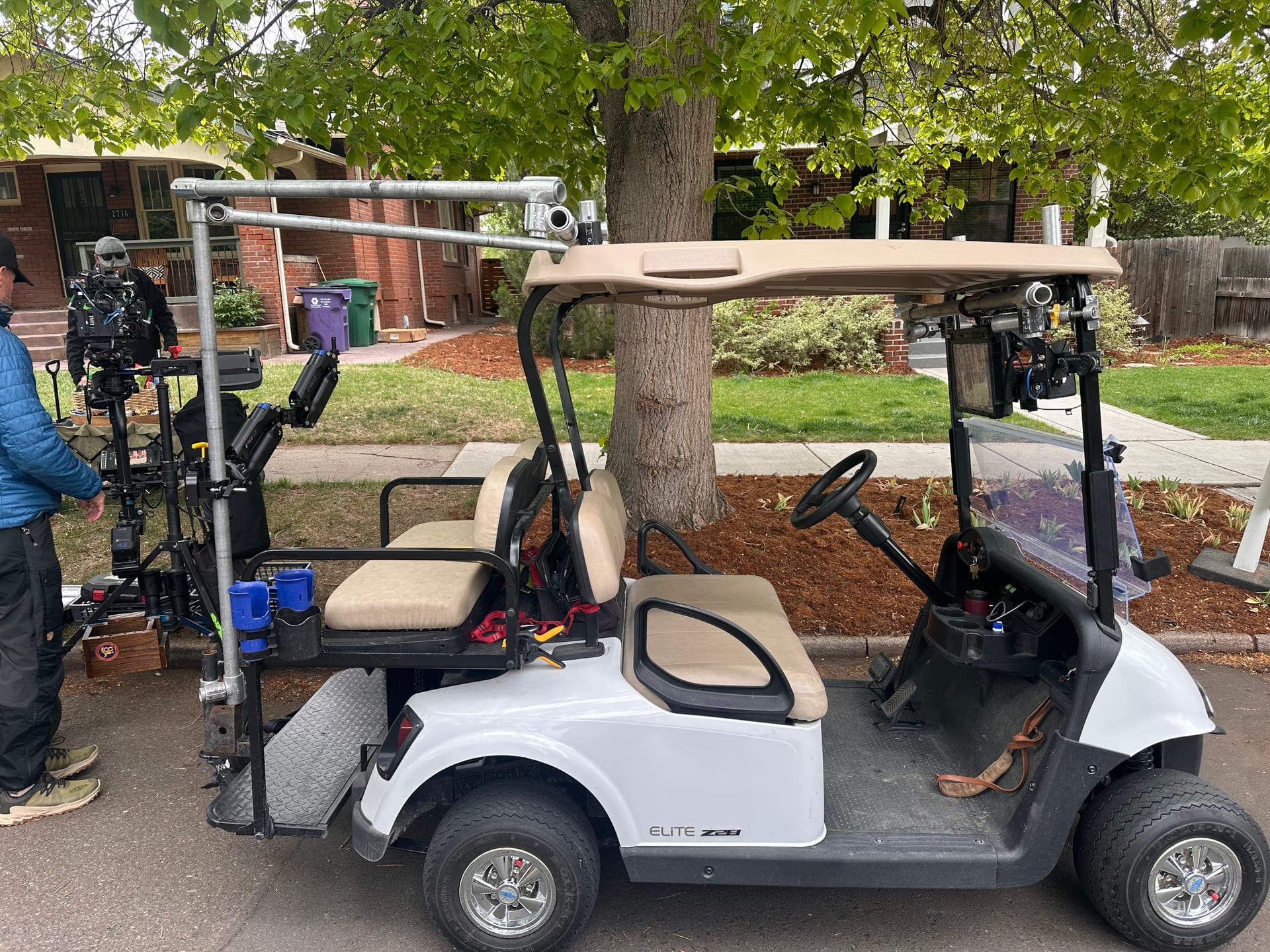 Modified white production golf cart rigged as a grip cart and camera car, with speed rail over the roof and a cinema camera on a rear-mounted gimbal; a crew member adjusts the rig on a suburban residential street.